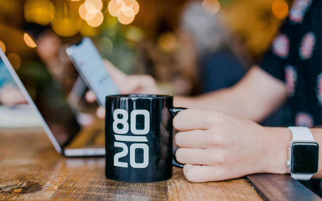 A person holds a mug with 80/20 printed on it, while working on a laptop.