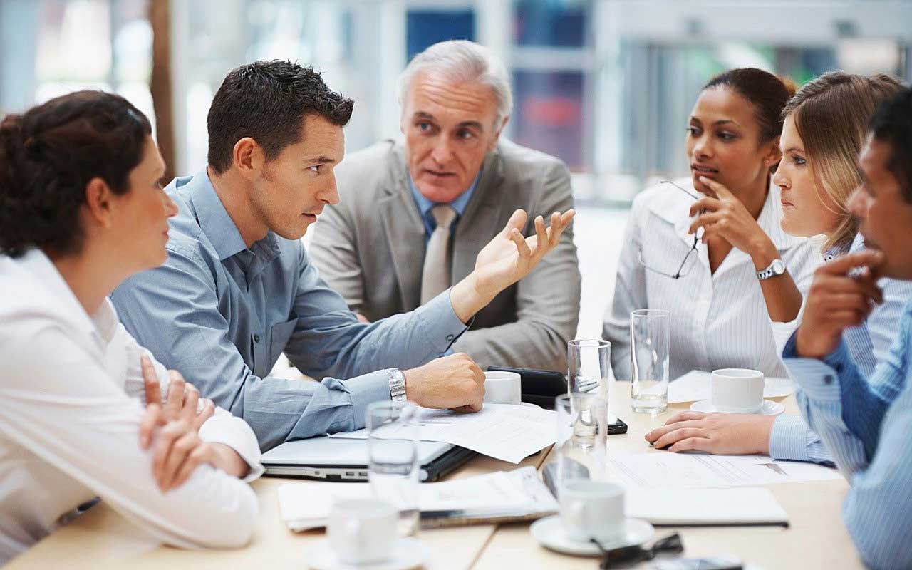 A racially and gender-diverse group of people dressed in business attire sit around a table, deep in discussion.