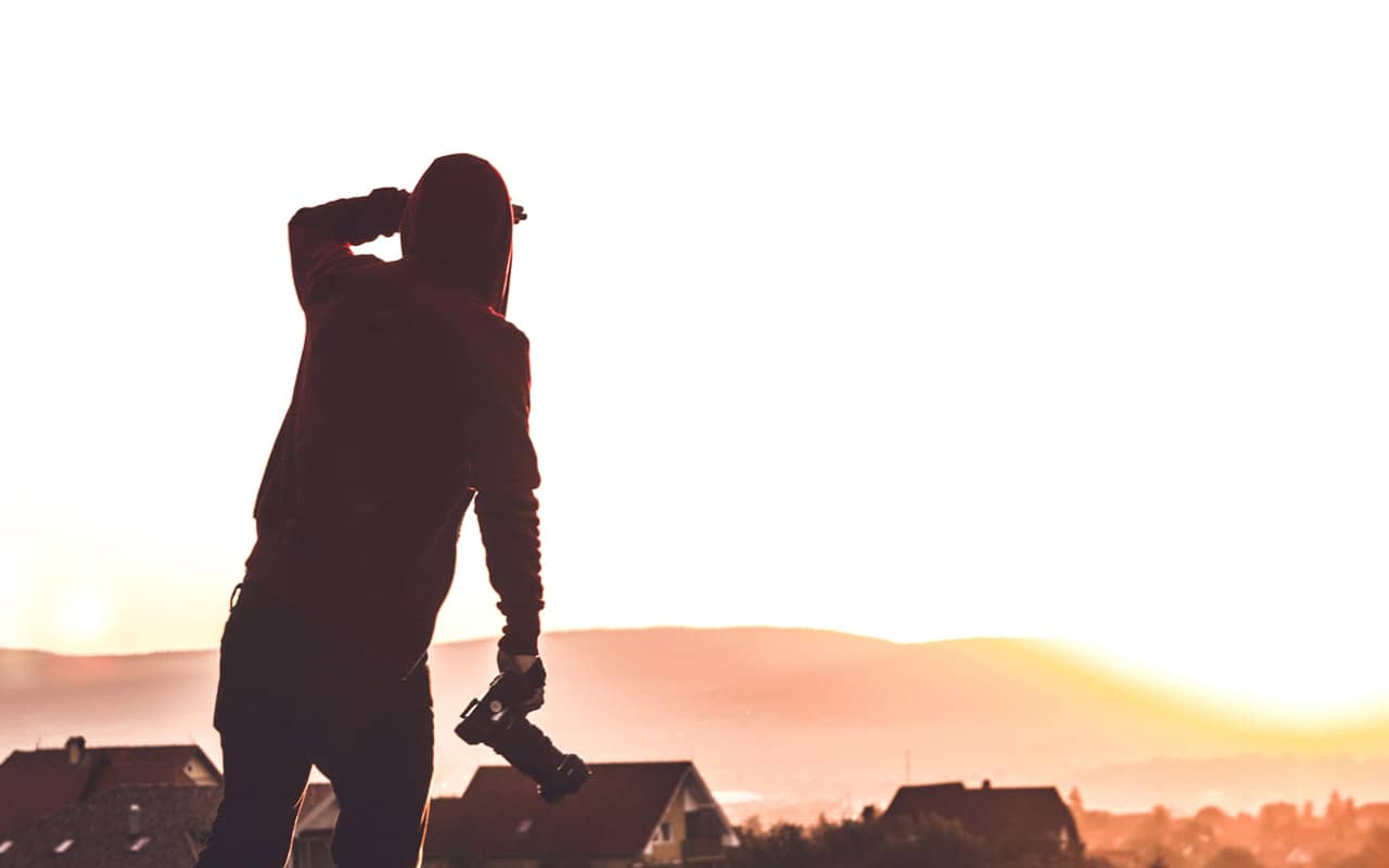 A man with a camera in his hand looks across a field during sunset.