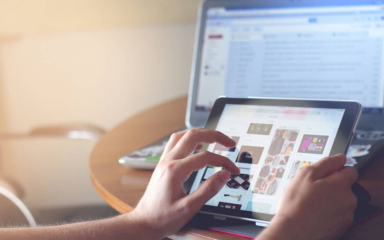 A person scrolls through a tablet while their laptop is open behind them. Limit your time mindlessly scrolling when you need to focus on work.