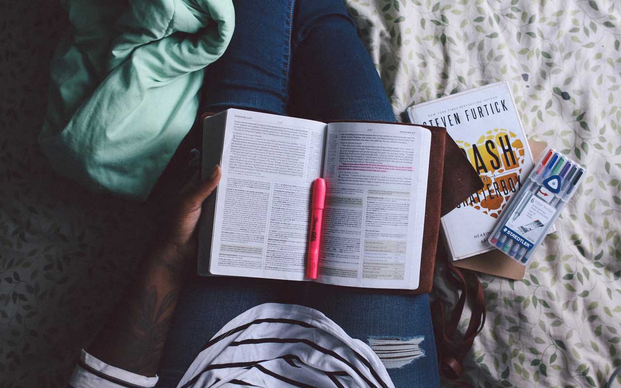A person sits on a patterned bedspread with a book and highlighter in their lap, as well as other books and pens next to them.
