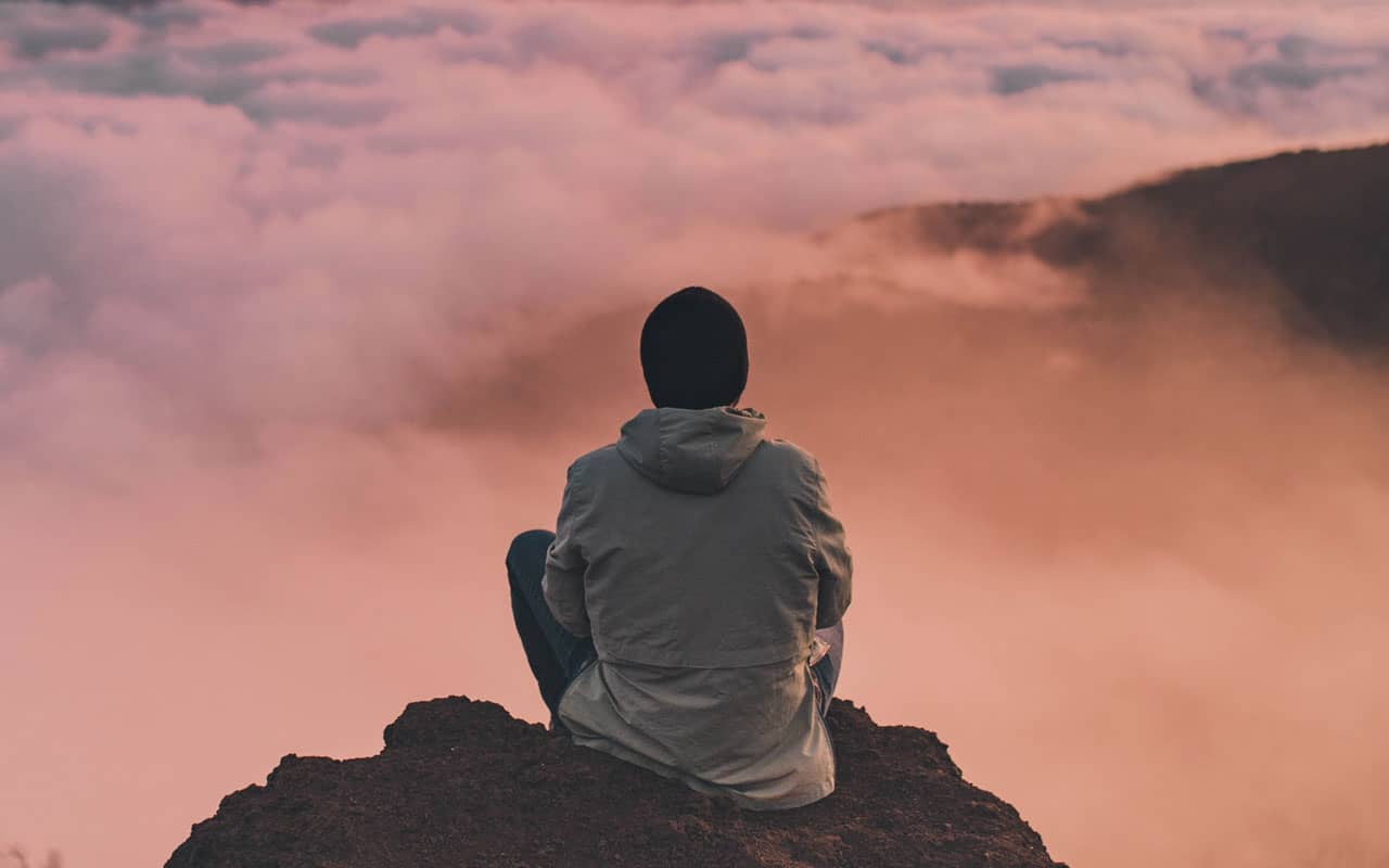 A person in a coat and winter hat sits at the top of a mountain at sunset, overlooking the clouds below.