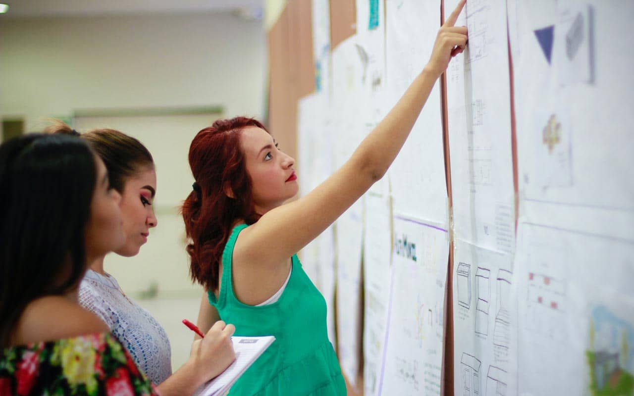 A woman in a green top points at papers on a bulletin board. A good teacher can help you learn Spanish faster.