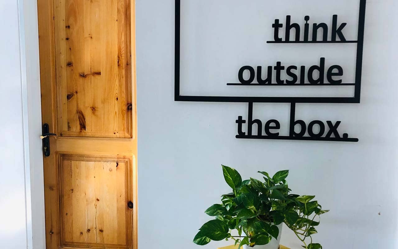 A sign with the text "think outside the box" against a white wall, with a green plant on a table underneath the sign.